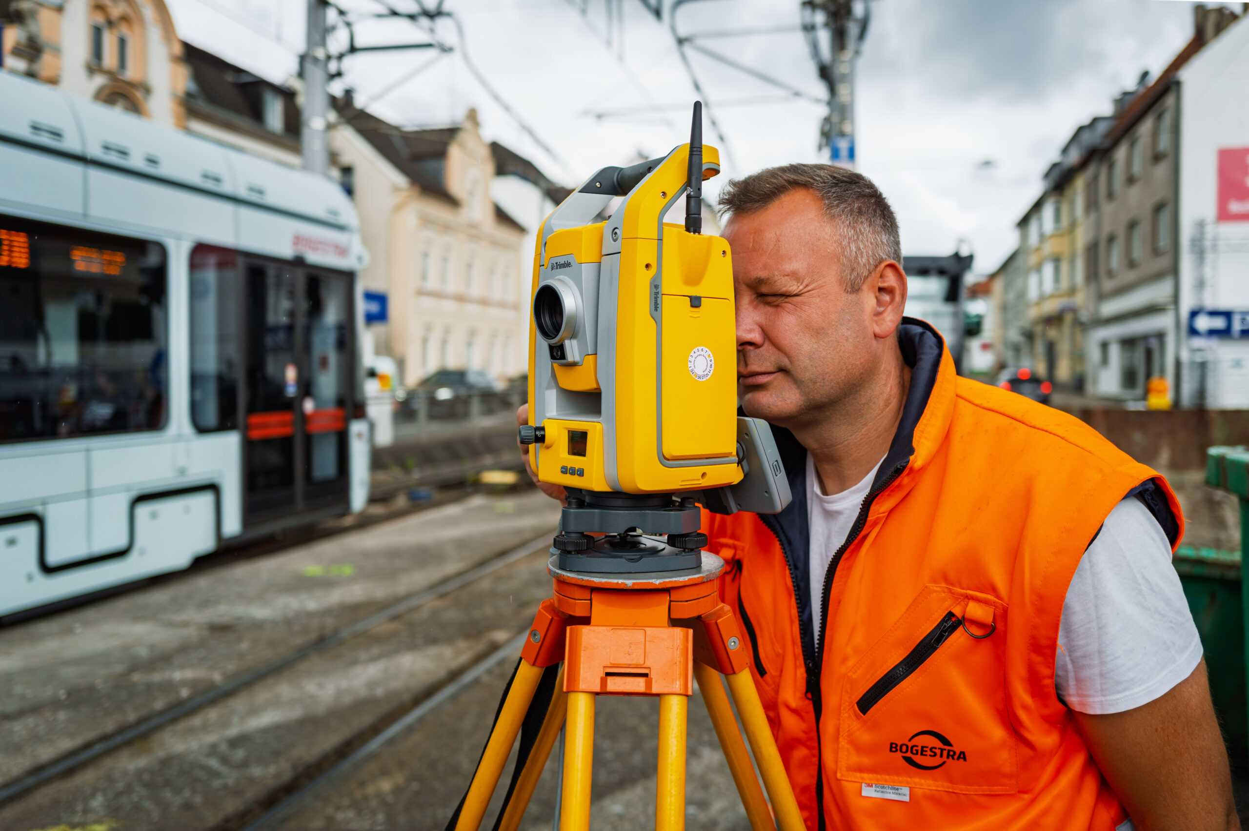 Christian guckt durchs Okular Vermessungsingenieur Christian guckt durch das Okular der Totalstation. Im Hintergrund fährt eine Straßenbahn vorbei.