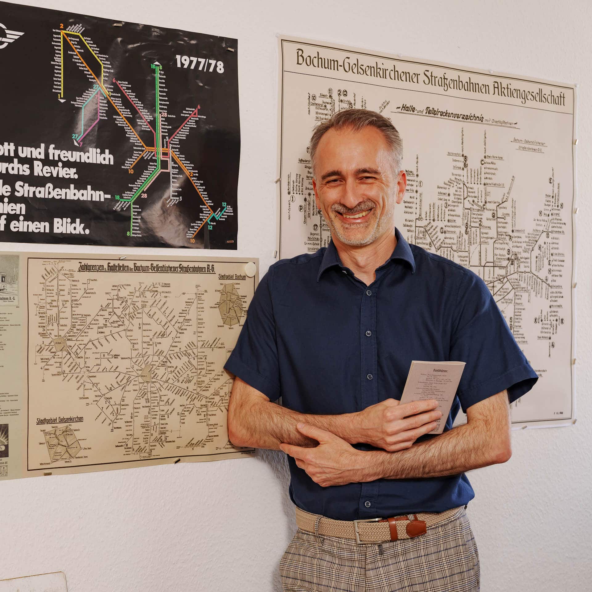 Unser Kollege Sven lehnt an der Wand seines Büros. Hinter ihm sind viele alte Liniennetzpläne.
